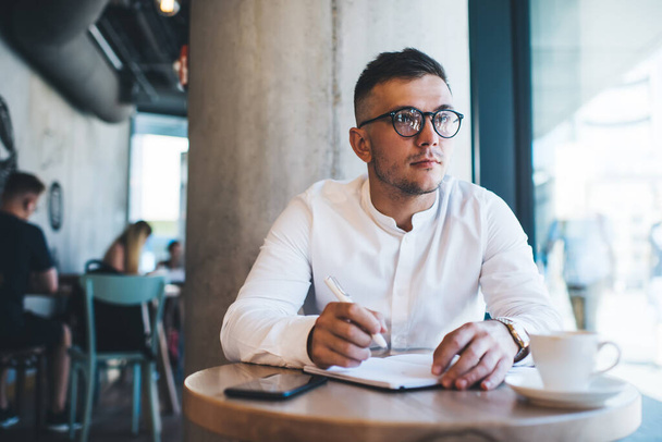 Young male in white shirt and eyeglasses taking notes with cup of hot drink while working in cafe terrace and looking away at daytime - 写真・画像