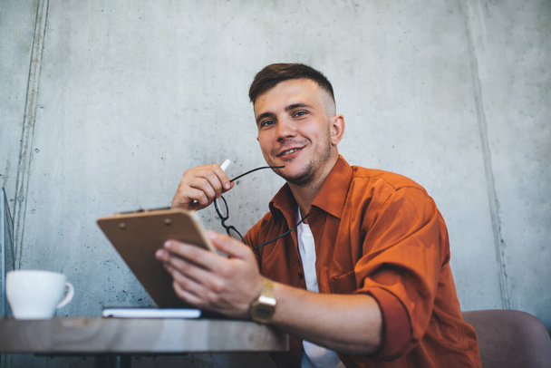 From below of joyful young male student in casual clothes studying notes on clipboard and smiling while sitting at table in cafe and looking at camera - Фото, зображення