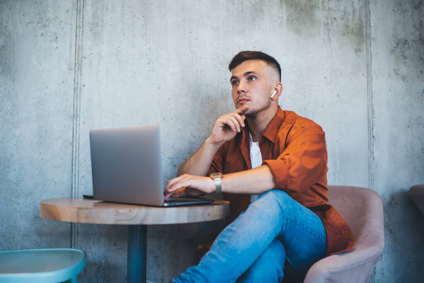 Сконцентрированный молодой человек в оранжевой рубашке и синих джинсах сидит за столом и печатает на ноутбуке, слушая музыку на беспроводных наушниках - Фото, изображение