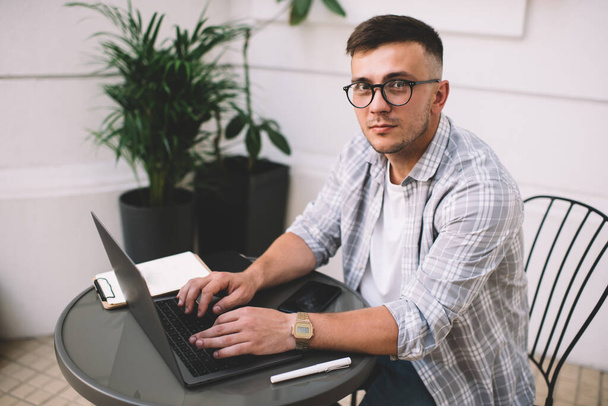 Positive smart freelancer in checkered shirt and eyewear browsing laptop while sitting at cozy workspace and attentively looking at camera - Fotó, kép