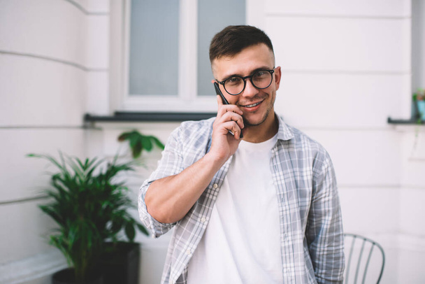 Positive bearded man in casual wear and eyeglasses smiling and looking at camera while standing in front of building with white striped wall - Foto, afbeelding