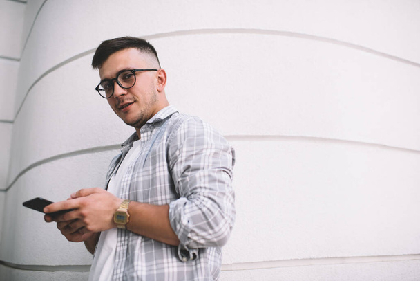 From below side view of young man in casually gray checkered shirt with wristwatch and smartphone standing near white wall and looking at camera - Fotó, kép