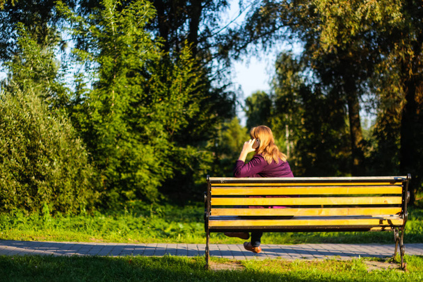 Defocus caucasian blond woman talking, speaking on the phone outside, outdoor. 40s years old woman in purple blouse in park on bench. Adult women using telephone. Back view. Copy space. Out of focus. - Photo, image