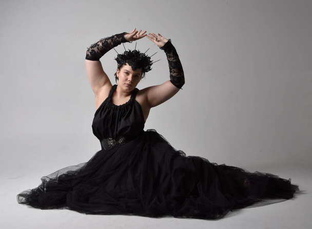 Full length portrait of young  plus sized woman with short  hair,  wearing long black tuille gothic gown with crown and gloves, sitting pose on light studio background. - 写真・画像