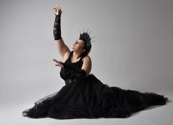 Full length portrait of young  plus sized woman with short  hair,  wearing long black tuille gothic gown with crown and gloves, sitting pose on light studio background. - Photo, image