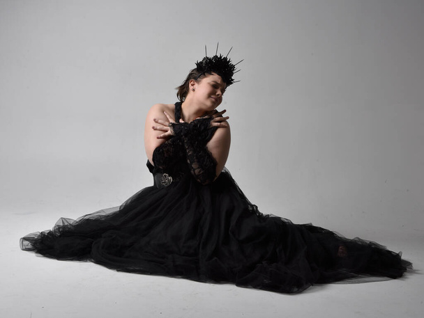 Ganzkörperporträt einer jungen Frau mit kurzen Haaren, die ein langes schwarzes Tuille-Gotikkleid mit Krone und Handschuhen trägt und auf hellem Studiohintergrund posiert. - Foto, Bild