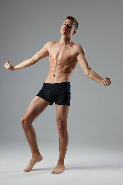 hombre desnudo en bragas negras sobre un fondo gris bombea los músculos del torso descalzo  - Foto, Imagen