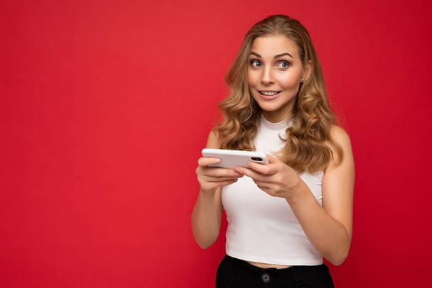 写真は、スマートフォンを使用して赤の背景に隔離された白いTシャツを身に着けている濃縮された感情的な美しい若いブロンドの女性の側を見て電話ゲームをプレイ - 写真・画像