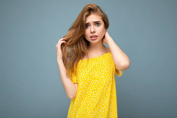 Молодая скорбящая грустная темноволосая блондинка с искренними эмоциями изолирована на задней стене с копировальным пространством в стильном летнем желтом платье. Грустная негативная концепция - Фото, изображение