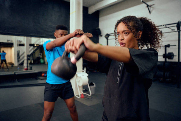 African American male personal trainer instructing African American female with a kettlebell routine in the gym. Mixed race friends doing cross training together. High quality photo - Photo, image