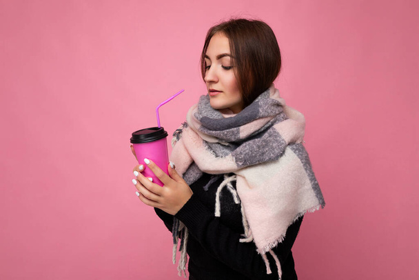 Foto shot van mooie jonge brunette vrouw dragen zwarte trui en warme sjaal geïsoleerd over roze achtergrond met papier koffiebeker voor mockup drinken en kijken naar de zijkant - Foto, afbeelding
