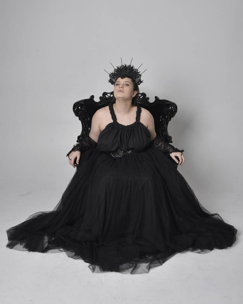 Full length portrait of young  plus sized woman with short  hair,  wearing long black tulle gothic gown with crown and gloves, sitting pose on a ornate throne chair on light studio background. - Fotografie, Obrázek