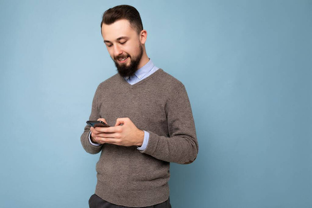 Photo shot of handsome positive good looking young man wearing casual stylish outfit poising isolated on background with empty space holding in hand and using mobile phone messaging sms looking at - Φωτογραφία, εικόνα
