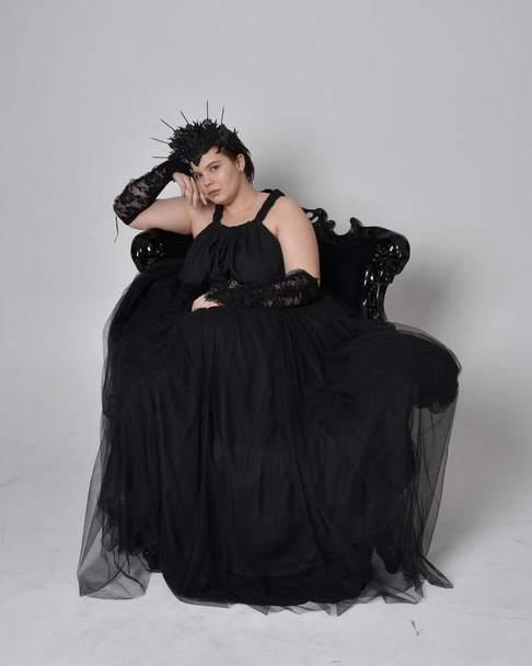 Full length portrait of young  plus sized woman with short  hair,  wearing long black tulle gothic gown with crown and gloves, sitting pose on a ornate throne chair on light studio background. - 写真・画像