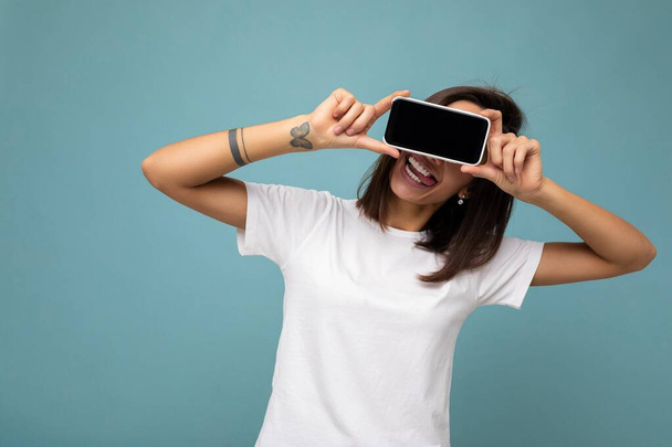 Photo of pretty positive adult brunette woman good looking wearing white t-shirt standing isolated on blue background with copy space holding smartphone showing phone in hand with empty screen screen - Φωτογραφία, εικόνα