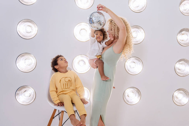 Young blond Caucasian woman holds an African American daughter in arms, son sits on stylish chair. Fashion family stands on a bright, textured background of led lamps, children play with a disco ball. - 写真・画像
