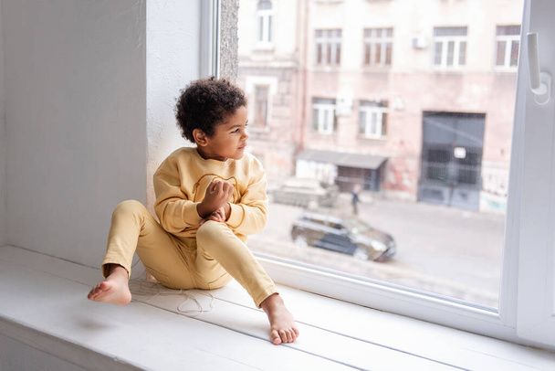A little African American boy in yellow pajamas sits barefoot on a wide windowsill by a large white window. The child is having fun, laughing and fooling around. Happy life style childhood at home. - Foto, afbeelding