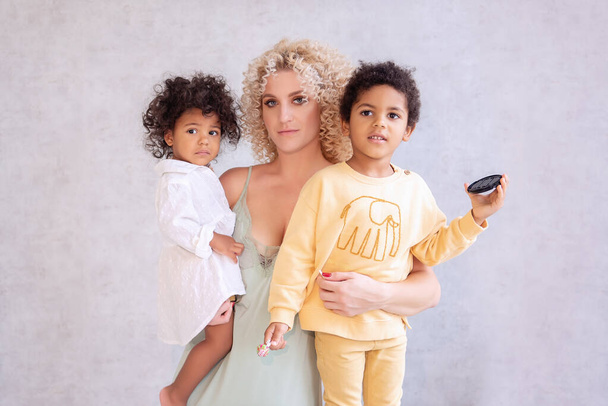 Young blond Caucasian woman holds in her arms, hugging, African American children, her daughter and son. Happy family stands on a bright, textured background of led lamps. Single Adoption Parenting. - Fotografie, Obrázek