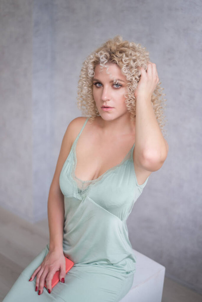 Close-up beauty portrait of caucasian young blonde woman with afro curls hairstyle with blue eyes. Girl in mint elegant dress on gray textured isolated background. Natural makeup, clear skin. - Foto, afbeelding