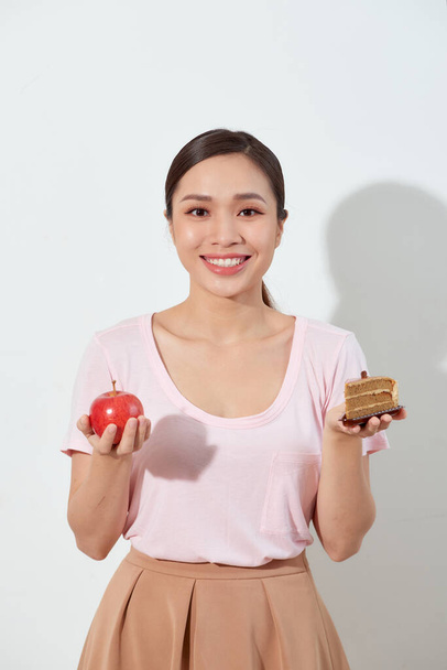 Portret zagubionej młodej Azjatki wybierającej między ciastem a jabłkiem, odizolowanej na białym tle. - Zdjęcie, obraz