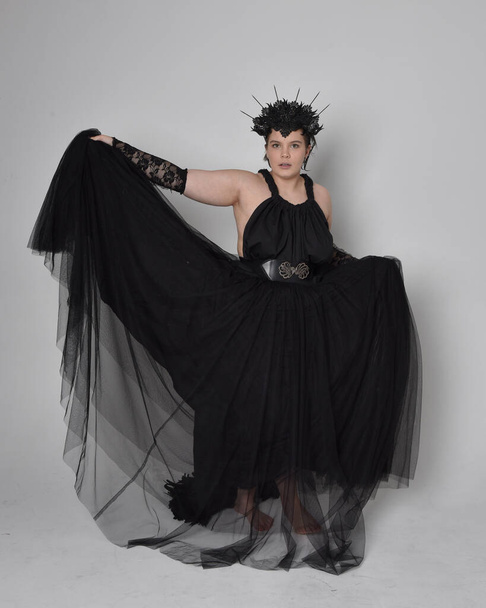 Full length portrait of young  plus sized woman with short  hair,  wearing long black tulle gothic gown with crown and gloves, standing pose on light studio background. - Foto, Imagem