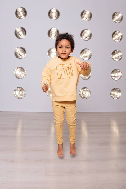 Little African American boy in yellow pajamas jumping barefoot on the laminate. White background with bright shades of led lamps. Stylish texture, trendy interior. Children's party in light style - Foto, Imagen