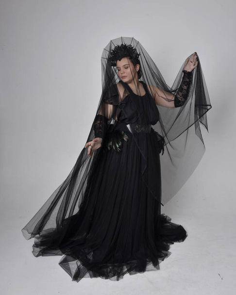 Full length portrait of young  plus sized woman with short  hair,  wearing long black tulle gothic gown with crow, gloves and veil,  standing pose on light studio background. - Foto, Bild