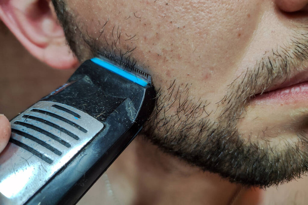 Muž si holí vousy elektrickým holicím strojem. Hezký mladý vousatý muž stříhání vousy s trimmer - Fotografie, Obrázek