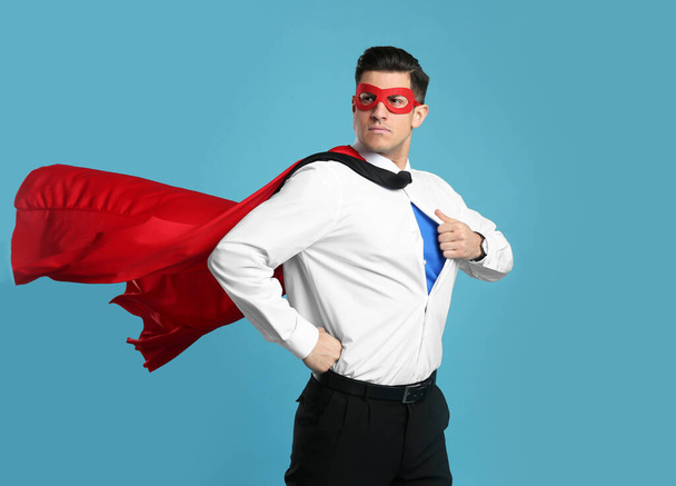 Businessman in superhero cape and mask taking shirt off on light blue background - Foto, afbeelding