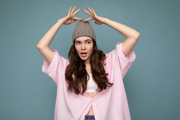 Foto der schönen emotionalen jungen brünetten Frau, die isoliert auf blauem Hintergrund Wand trägt rosa Hemd und grauen Hut, der zur Seite schaut - Foto, Bild