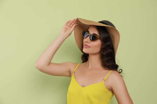 Beautiful young woman with straw hat and stylish sunglasses on light green background - Fotó, kép