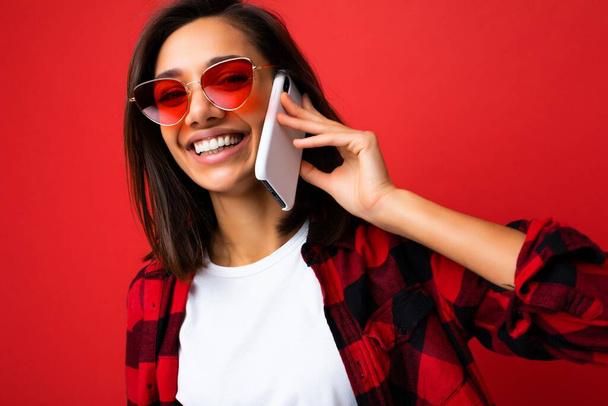Κοντινή φωτογραφία μιας πολύ χαρούμενης νεαρής μελαχρινής γυναίκας με κομψό κόκκινο πουκάμισο λευκό μπλουζάκι και κόκκινα γυαλιά ηλίου που απομονώνονται σε κόκκινο φόντο και επικοινωνούν στο κινητό τηλέφωνο κοιτάζοντας την κάμερα - Φωτογραφία, εικόνα