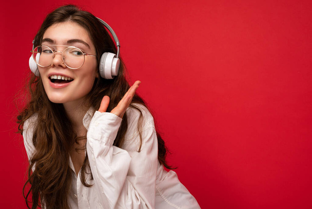 Fénykép gyönyörű boldog mosolygós fiatal barna nő visel fehér ing és optikai szemüveg elszigetelt felett piros háttér fal visel fehér vezeték nélküli bluetooth fejhallgató hallgat hűvös zene - Fotó, kép