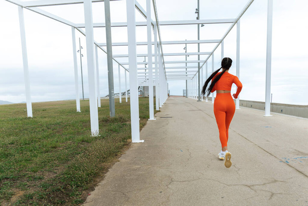 Modern şehir parkında koşan bayan sporcu. Neon turuncu spor giysili kadın koşucu parkın yakınında çimentoda koşuyor. Aktif yaşam tarzı konsepti. Sağlık konsepti. Boşluğu kopyala - Fotoğraf, Görsel