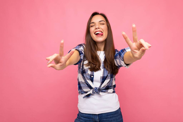 Young positive delightful smiling pretty brunette woman with sincere emotions wearing hipster check shirt standing isolated on pink background with free space and showing peace sign - Foto, afbeelding