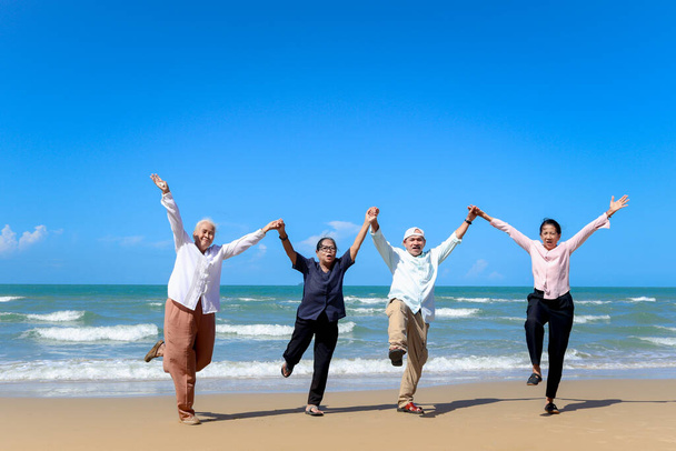 Grupa czterech wesołych starszych przyjaciół podróżuje razem na świeżym powietrzu, trzymając się i podnosząc ręce stojąc na plaży, bawiąc się i ciesząc spędzając czas, odpoczywając i relaksując na wakacjach.  - Zdjęcie, obraz