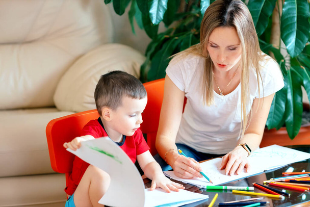 Uma bela jovem mãe de aparência caucasiana desenha lápis coloridos com seu filho no álbum. - Foto, Imagem
