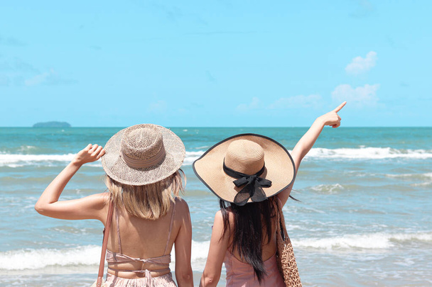 Dos hermosas mujeres asiáticas con sombrero grande por detrás disfrutan pasar tiempo en la playa de arena tropical mar azul juntos, descansando y relajándose en las vacaciones de verano. - Foto, Imagen