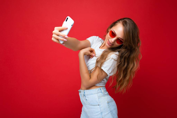 sexy piękny młody blondynka kobieta gospodarstwa telefon komórkowy biorąc selfie zdjęcie za pomocą smartfona aparat noszenie okulary przeciwsłoneczne codziennie stylowy strój odizolowany nad kolorowe tło ściany patrząc na urządzenie - Zdjęcie, obraz