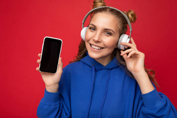Fénykép gyönyörű mosolygós fiatal nő visel stílusos kék kapucnis elszigetelt piros háttér visel fehér bluetooth vezeték nélküli fejhallgató és zenét hallgatni, és mutatja a mobiltelefon üres - Fotó, kép