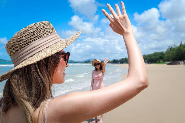 Schöne asiatische Frauen mit großem Hut und Sonnenbrille heben die Hand, um Freund am tropischen Sandstrand blaues Meer zu begrüßen, glückliche weibliche Freundin treffen und Gruß im Sommerurlaub. - Foto, Bild