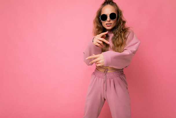 Photo shot of beautiful happy young dark blonde woman wearing casual clothes and stylish sunglasses isolated over colorful background looking at camera and having fun - Фото, изображение