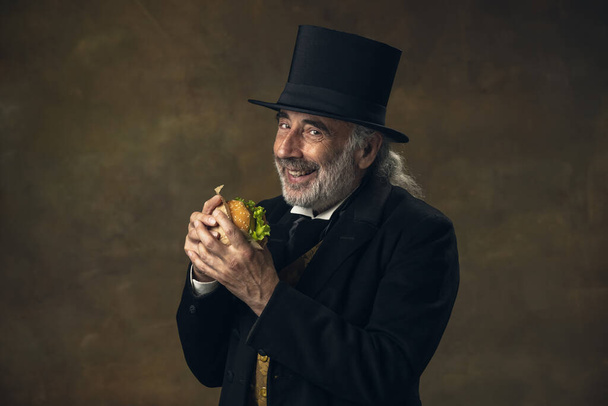 Handsome elderly gray-haired man, gentleman, aristocrat or actor eating fast food isolated on dark vintage background. Retro style, comparison of eras concept. - Photo, image