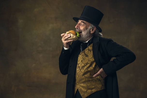 Handsome elderly gray-haired man, gentleman, aristocrat or actor eating fast food isolated on dark vintage background. Retro style, comparison of eras concept. - Foto, afbeelding