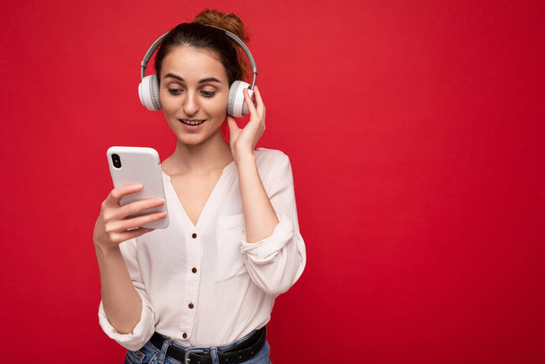 Photo shot of beautiful joyful smiling young female person wearing stylish casual outfit isolated over colorful background wall wearing white bluetooth wireless earphones and listening to music and - Zdjęcie, obraz