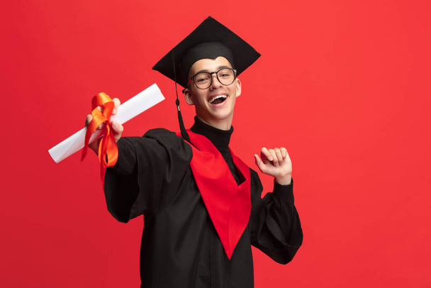 Young smiling, cheerful student, graduate in mortarboard with scroll isolated over red studio background. - Foto, Bild