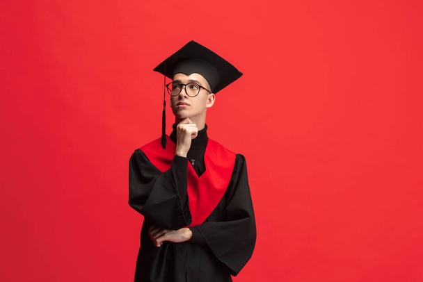 Young caucasian guy, student, graduate in mantle and mortarboard isolated over red studio background. - Foto, immagini