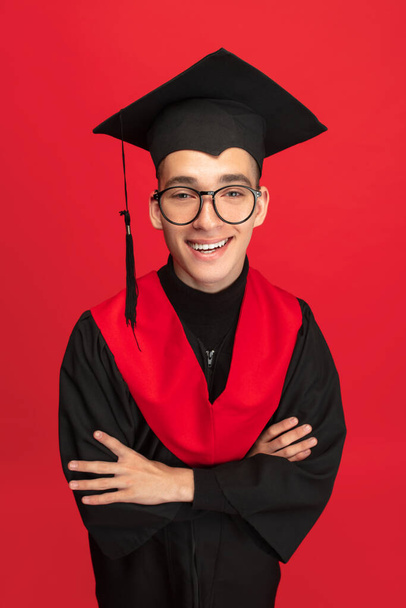 Studio shot of young smiling, cheerful student, graduate in mortarboard isolated over red studio background. - Photo, Image