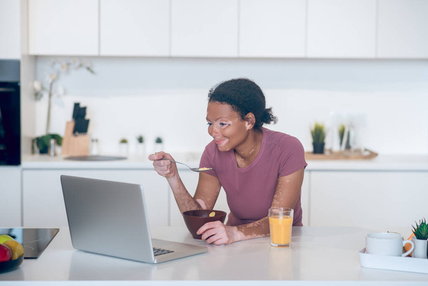 Sötét bőrű fiatal nő otthon reggelizik és néz valamit egy laptopon - Fotó, kép
