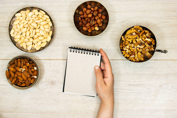 Assortment of nuts in metal bowls hazelnuts, walnuts, almonds and hand holding a empty notebook - Photo, Image